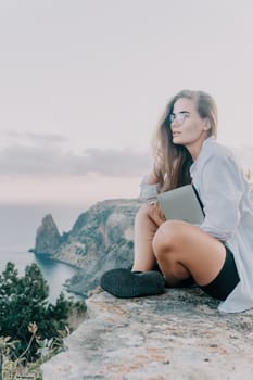 Digital nomad, woman in the hat, a business woman with a laptop sits on the rocks by the sea during sunset, makes a business transaction online from a distance. Freelance, remote work on vacation.