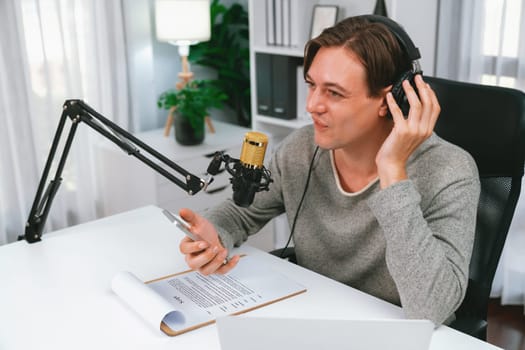 Host channel in smiling smart broadcaster recording, talking show with script and smartphone on desk on live social media, wearing headphones to record video streamer at modern home studio. Pecuniary.