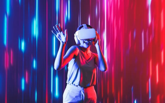 Smart female standing surrounded by neon light wearing VR headset connecting metaverse, future cyberspace community technology. Elegant woman enjoy dancing wavering body and sing song. Hallucination.