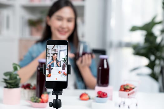 Selective focus smartphone with smiling young beautiful Asian reviewing mixed strawberry and cherry juice detox drink, presenting healthy product promotion selling on social media online. Stratagem.