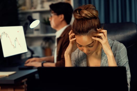 Businesswoman working on desk with stretching arm up and down manner with body health ache of strain overwork while coworker trading stock market on website on pc at late over night time. Infobahn.