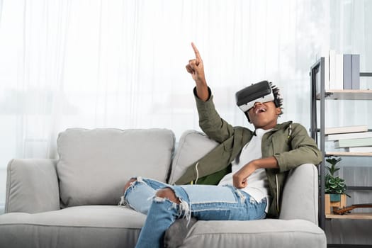 Surprised African American looking VR enjoy watching by touching the air in fantastic 3D cartoon movie technology virtual reality metaverse journey new world at living room sitting sofa. Contrivance.