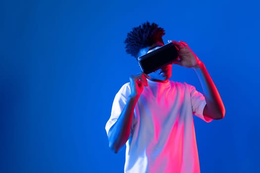 Surprised young African American looking through VR holding interesting object hologram on pink blue neon wall at metaverse world connecting digital futuristic technology virtual reality. Contrivance.