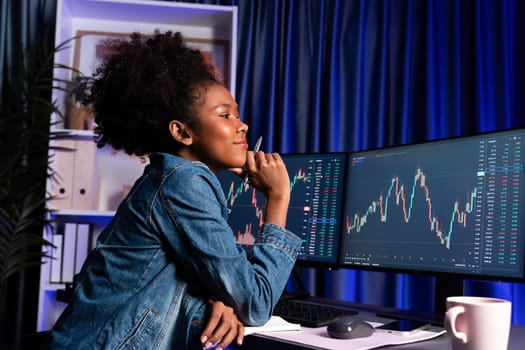 Young African woman trader smiling on happy face, looking on screen with valued stock market chart at high profit. Concept of exchange investment online in trading application on pc. Tastemaker.