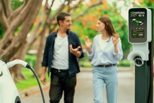 Focused EV car recharging battery on blurred background of lovey couple during autumnal road trip travel with electric vehicle recharging battery. Eco friendly travel on vacation during autumn. Exalt