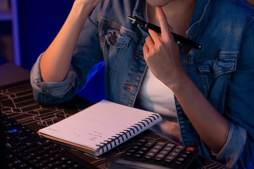 Cropped image of young Asian creative woman calling on phone to customer or coworker for marketing creator plan on pc with notebook and pen to write memo at neon modern office at night. Stratagem.