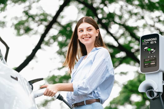 Young woman recharging battery for electric car during road trip travel EV car in natural forest or national park. Eco friendly travel during vacation and holiday. Exalt