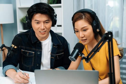 Smiling radio influencer hosts wearing headphones with channel social media online live streaming with talking topic on script note and laptop to listeners at morning time at studio record. Infobahn.