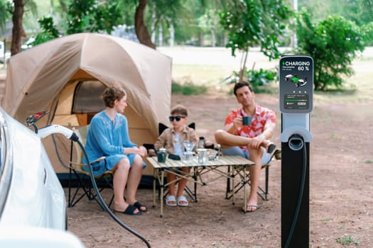 Outdoor adventure and family vacation camping in nature travel by eco friendly car for sustainable future. Lovely sit on trunk, charging EV car with EV charging station in campsite. Perpetual