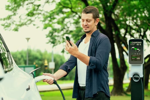 Man using smartphone online banking application to pay for electric car battery charging from EV charging station during vacation holiday road trip at national park or summer forest. Exalt