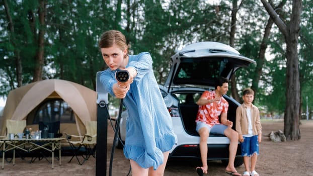 Outdoor adventure and family vacation camping at sea travel by eco friendly car. Cheerful woman or mother holding, pointing EV charger point with playful and happiness posture in campsite. Perpetual