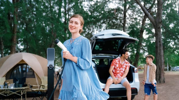 Outdoor adventure and family vacation camping at sea travel by eco friendly car. Cheerful woman or mother holding, pointing EV charger point with playful and happiness posture in campsite. Perpetual