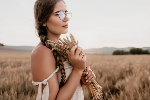 A woman is holding a bunch of wheat in her arms. The wheat is dry and brown, and the woman is wearing a white dress. The scene is set in a field, and the woman is posing for a photo