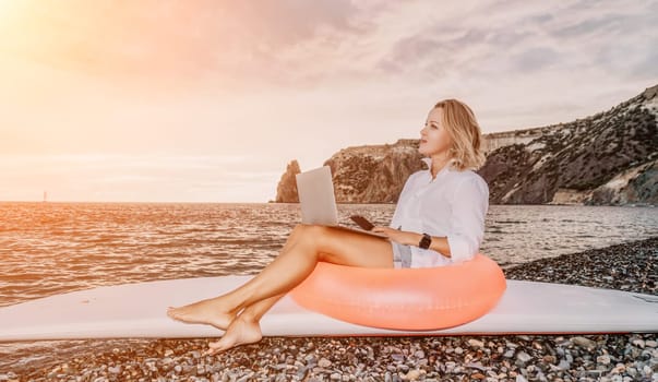 Successful business woman in yellow hat working on laptop by the sea. Pretty lady typing on computer at summer day outdoors. Freelance, travel and holidays concept.