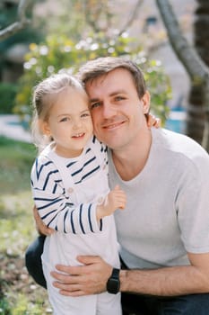 Little girl hugs the neck of her smiling dad squatting in the garden. High quality photo