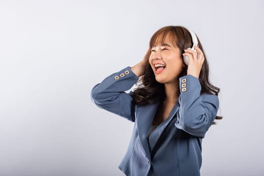 Happy Asian young female in Bluetooth headphones, smiling and listening to music on her phone. Studio shot, isolated on white background. She's embracing modern technology with joy.