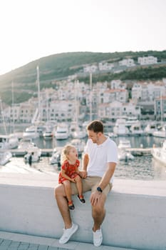 Little laughing girl sits on her dad lap on the fence of the luxury marina embankment. High quality photo