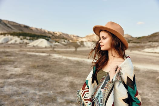 Woman in hat and blanket standing alone in desert with mountains in background on cloudy day