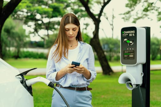 Young woman recharge EV electric vehicle battery from EV charging station and using smartphone online banking to pay for electricity in city park. Eco friendly vehicle travel with EV car. Exalt