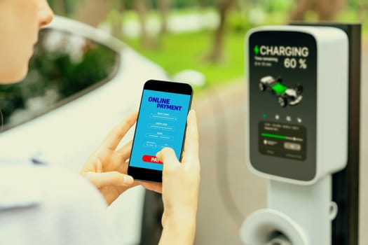 Young woman using smartphone online banking application to pay for electric car battery charging from EV charging station during vacation holiday road trip at national park or summer forest. Exalt