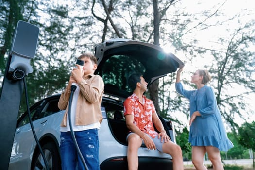 Little boy holding EV charger and point at camera with his family sitting on the trunk in background. Road trip travel with alternative energy charging station for eco-friendly car concept. Perpetual