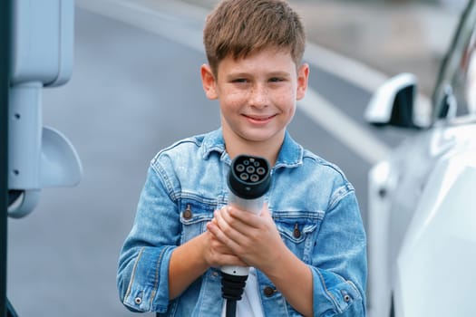 Road trip vacation with eco-friendly EV car and little boy pointing EV charger at camera. Electric vehicle and charging station using clean and sustainable energy for environment protection. Perpetual