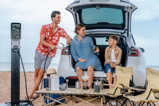 Family vacation trip traveling by the beach with electric car, happy family recharge EV car, enjoying outdoor camping coffee. Seascape travel and eco-friendly car for clean environment. Perpetual