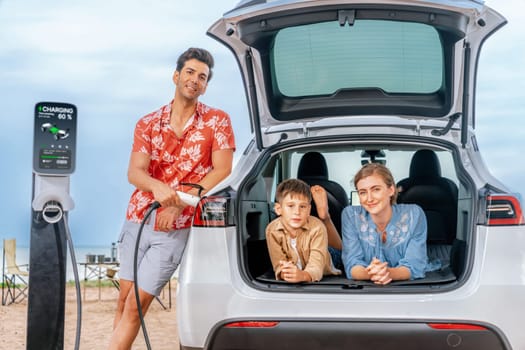 Family vacation trip traveling by the beach with electric car, lovely family sit on the trunk, charging EV car battery with green and sustainable energy. Family travel and eco-friendly car. Perpetual