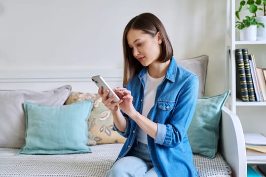 Young woman using smartphone sitting on couch at home. Female texting reading looking, mobile applications technology internet online services for work study leisure communication shopping lifestyle