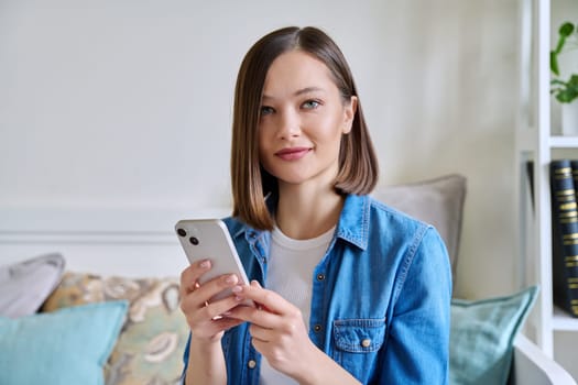 Young smiling woman using smartphone sitting on couch at home. Female looking at camera texting reading looking mobile applications technology internet online services for work study leisure lifestyle