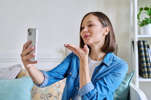 Young woman sitting with smartphone on couch at home having video call. Smiling female blowing kiss to the camera, chatting with friends colleagues on social networks