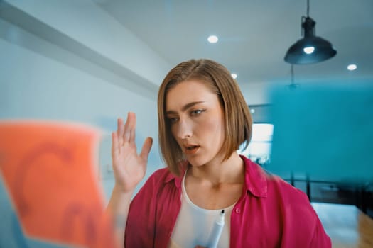 Portrait of young caucasian businesswoman thinking with confused face expression while standing in front of glass board with sticky notes and mind map at creative business meeting. Immaculate.