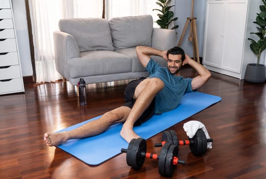 Athletic and sporty man doing crunch on fitness mat during home body workout exercise session for fit physique and healthy sport lifestyle at home. Gaiety home exercise workout training concept.