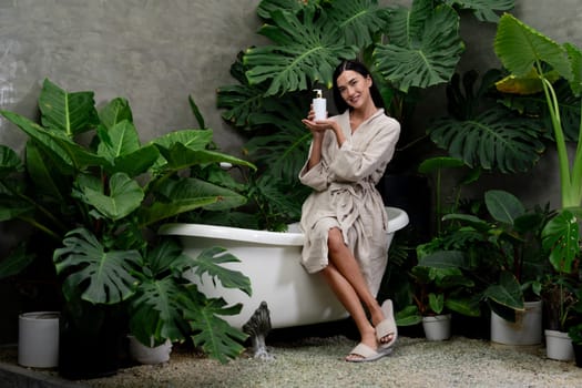 Tropical and exotic spa garden with bathtub in modern hotel or resort with woman in bathrobe holding beauty skincare product while enjoying leisure lush with greenery foliage background. Blithe