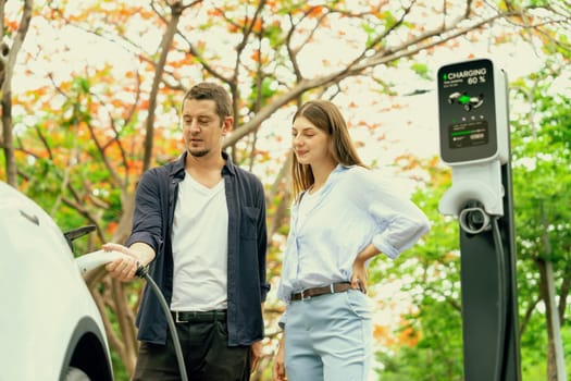 Lovely young couple recharging battery for electric car during autumnal road trip travel EV car in autumnal forest. Eco friendly travel on vacation during autumn. Exalt