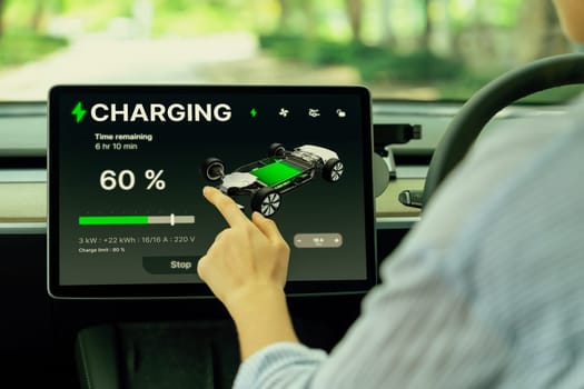 Young woman inside the vehicle, checking EV vehicle's application on battery recharging electricity status display on smart monitor screen in modern EV car on her road trip journey. Exalt