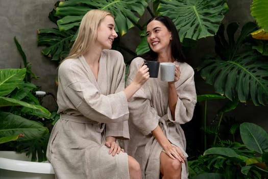 Tropical and exotic spa garden with bathtub in modern hotel or resort with young two women in bathrobe drink coffee, enjoy leisure and wellness lifestyle surround by lush greenery foliage. Blithe