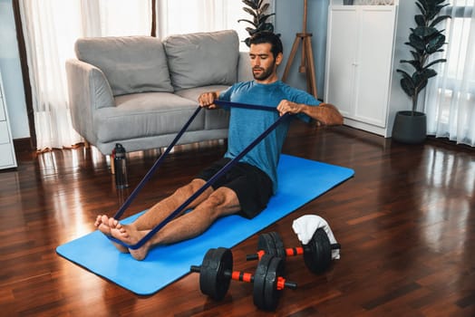 Athletic and sporty man pulling resistance band exercise during home workout exercise session for fit physique and healthy sport lifestyle at home. Gaiety home exercise workout training concept.