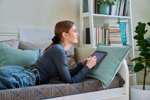 Young female teenager with digital tablet, stylus drawing illustrating lying on couch at home. Technologies, leisure, creativity, education, freelance work, lifestyle, youth concept