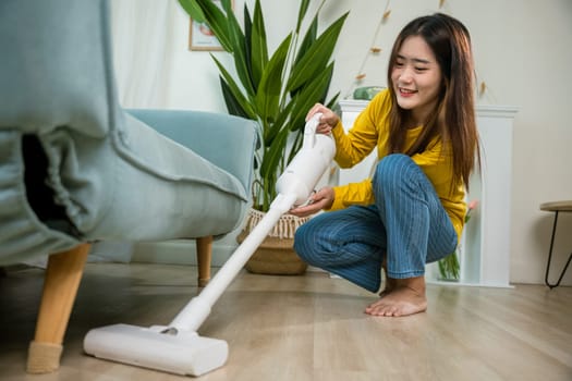 Housewife female dust cleaning floor under sofa or couch furniture with vacuum cleaner, Happy Asian young woman with accumulator vacuum cleaner at home in living room, household and housework concept