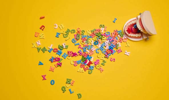 Artificial jaw with teeth, from which multi-colored letters of the alphabet spilled out on a yellow background, top view