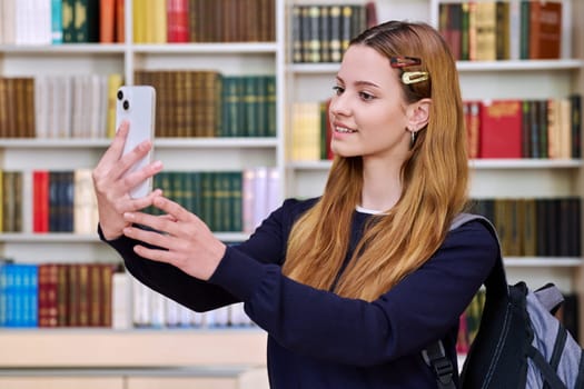 Teenage girl high school student taking selfie photo on smartphone inside library classroom. Education, technology, adolescence concept