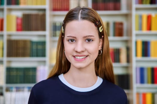 Headshot portrait of smiling teenage girl student. Attractive female teenager 16,17 years old inside high school building, background of classroom library shelves books. Education, adolescence
