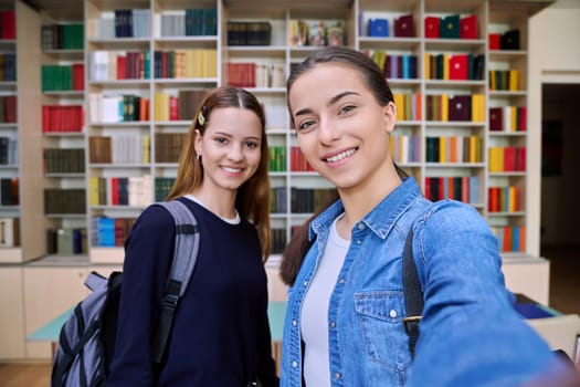 Selfie portrait of happy friends teenagers high school students looking at camera inside classroom. Smiling cheerful two teenage girls in library. Friendship adolescence education lifestyle concept