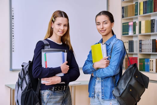 Portrait of two teenage female students together in library classroom. Smiling teenage girls classmates with backpacks textbooks looking at camera. High school education adolescence friendship concept