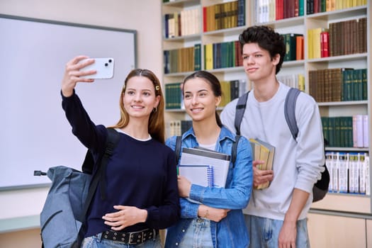 Selfie portrait of group friends teenagers high school students looking at camera inside classroom. Having fun cheerful teenage guy and two girls in library. Friendship adolescence education lifestyle