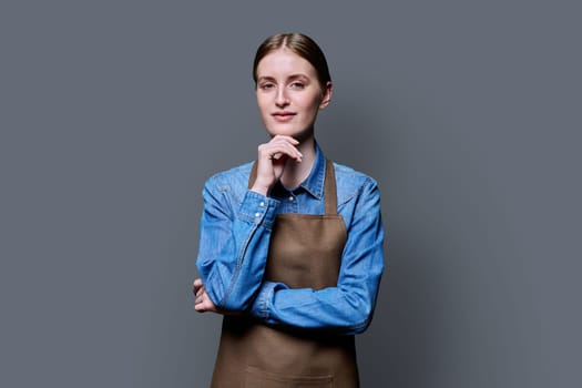 Portrait of young smiling confident woman in apron on grey studio background. Successful positive female looking at camera. Worker, startup, small business, service sector, staff, youth concept