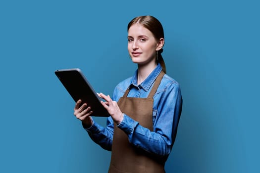 Young smiling female worker in apron uniform holding digital tablet on blue studio background. Digital technologies in business work, Internet apps applications for online orders and customer service