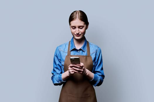 Young female worker in an apron using smartphone on gray studio background. Work, business, online internet services, mobile apps applications, technology concept
