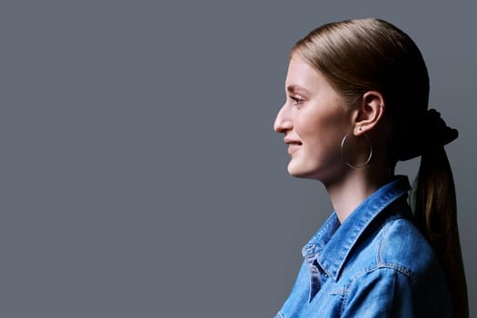 Profile view, young smiling female looking forward on gray studio background, copy space for advertising text image. Beautiful teenage blonde with long hair in denim shirt, fashion style beauty youth